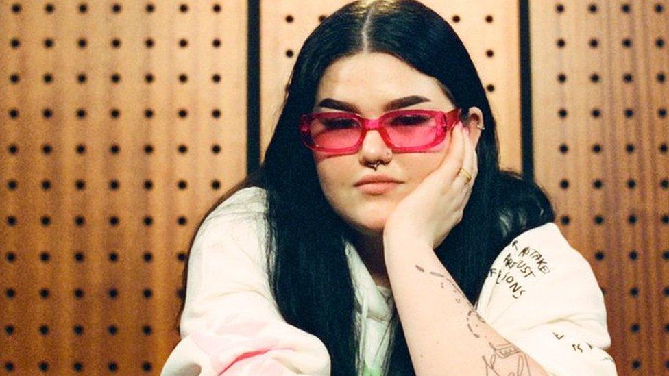 Woman with long dark hair and wearing white top with red glasses sits with her chin in her palm