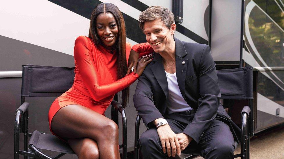 AJ Odudu, wearing a red dress, and Big Brother co-host Will Best, wearing a black suit, sit in black director-style chairs, smiling