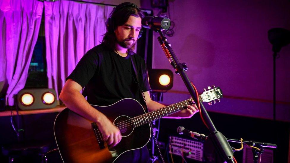 Noah Kahan in the BBC Radio 1 Live Lounge. Noah, 26, has brown hair down to his shoulders and a short brown beard. He wears a black T-shirt and is playing an acoustic guitar. The live lounge is lit by a purple light and behind him you can see sound equipment, lights and curtains. He stands in front of a microphone.