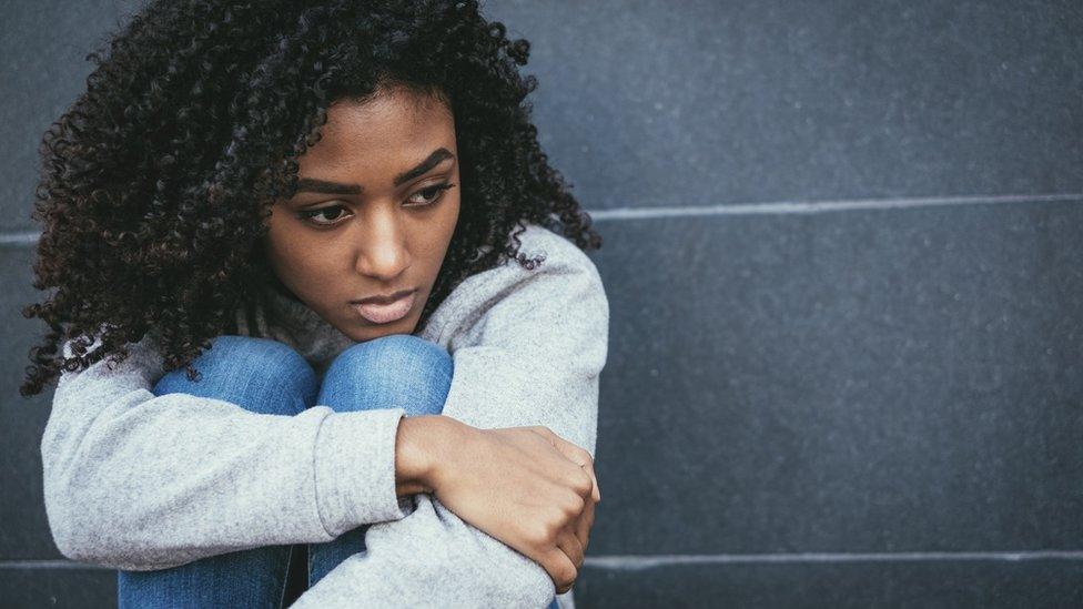 A woman sits against a grey wall