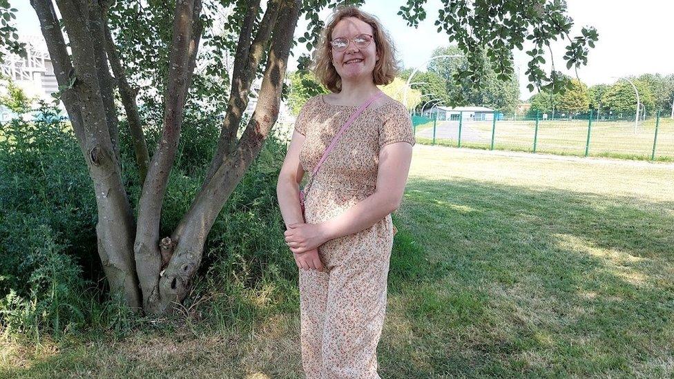 Rebecca is standing in a park, she is smiling at the camera and wearing a beige dress with flowers on