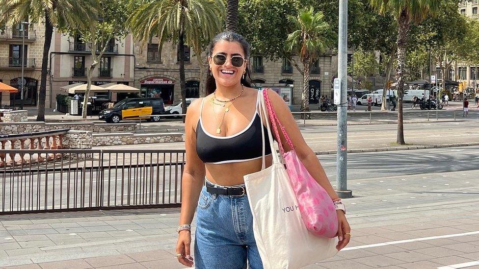 Rosie Breen on holiday standing and smiling for the camera. She has her brunette hair pulled back in a ponytail and is wearing black sunglasses. She is wearing earrings in both ears and has a few necklaces around her neck. She is wearing a black cropped vest top and blue denim shorts. She has two bags over her left shoulder, one is white and one is pink. Behind her there is a road and some trees before there is a row of buildings