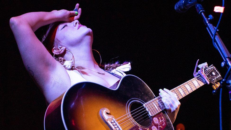 Singer CMAT on-stage, her head is thrown back and her eyes are closed. She has the back of one hand pressed to her forehead and the other resting on the fretboard of her acoustic guitar