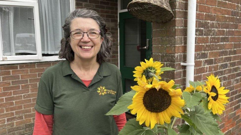 A woman smiling to the camera wearing a dark green top and standing next to some sunflowers.