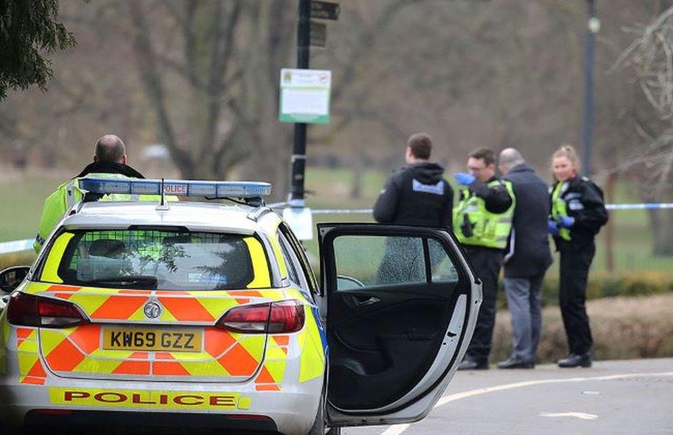 A police car in the foreground with officers and police tape in the background next to a grassy area of a park