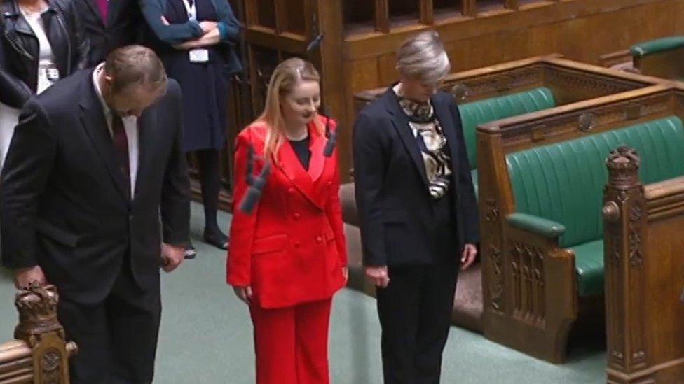 Woman in red, flanked by two other MPs, bows at a thick white line on the floor of the House of Commons