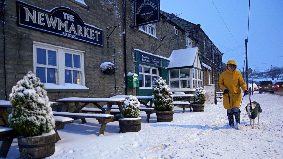 A woman in a yellow coat walks a dog along a snow covered path next to a pub