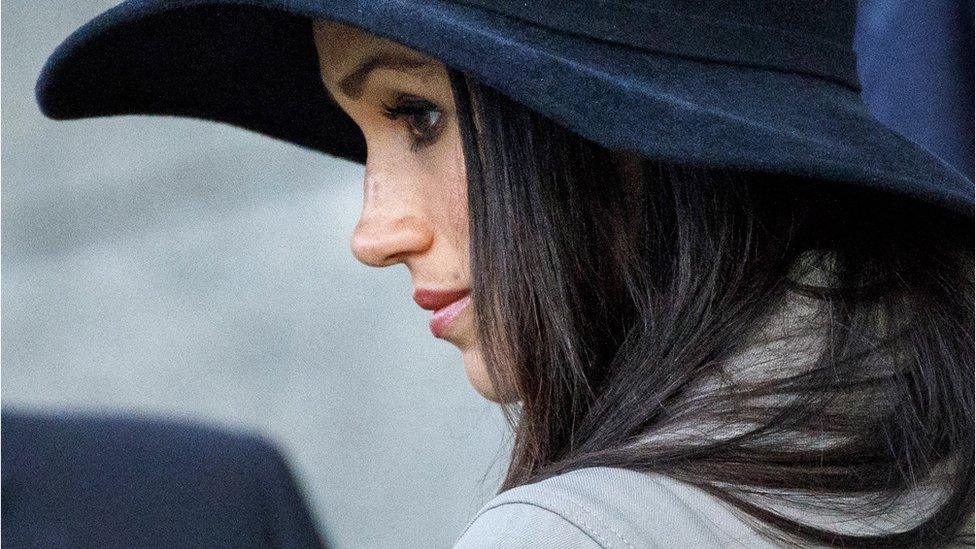 Image shows Meghan looking to her left wearing a black hat.