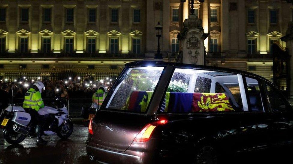 The Queen's coffin in a hearse, driving past Buckingham Palace with crowds and security nearby.