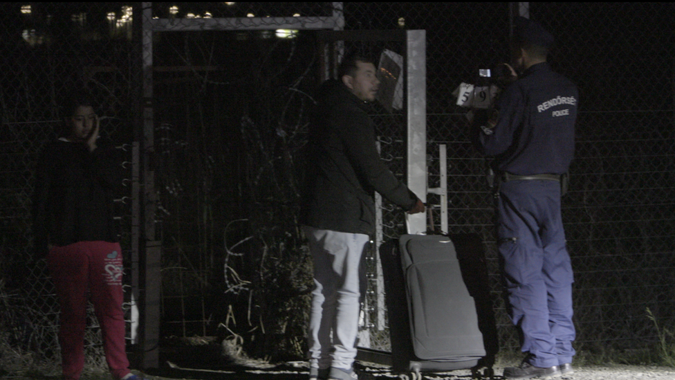 A man carrying a suitcase is photographed next to a woman, both of whom are being accompanied by a uniformed police officer nearby