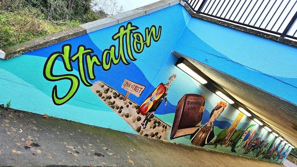 An underpass with a mural on one side, with a background in different shades of blue with paintings of a Roman soldier, the Domesday book and a church