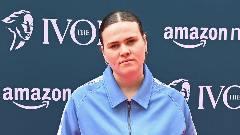 Venbee, standing in front of a red carpet advert hoarding showing the logo of the Ivor Novello Awards. She wears a light blue tracksuit top, her hair is tied back in a tight ponytail.