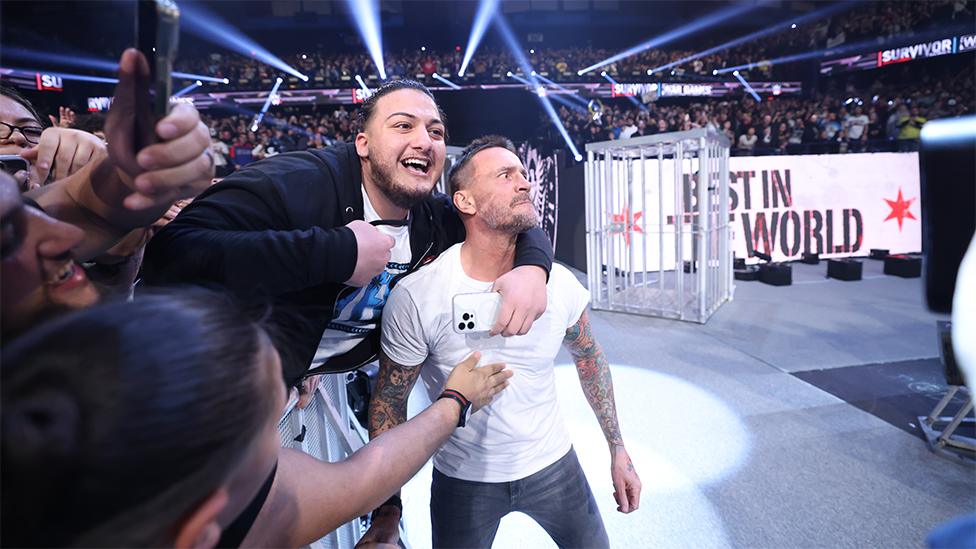 CM Punk at Survivor Series, with fans holding onto him looking really excited. Punk is wearing a white tshirt with his tattoos on his arms visible, with the fan wearing a tshirt and black top. The background has a screen which reads "best in the world", with flashing lights.