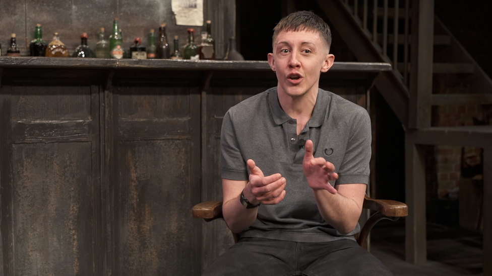 Charlie Josephine, who wrote the play, is sitting giving an interview on stage in a western bar