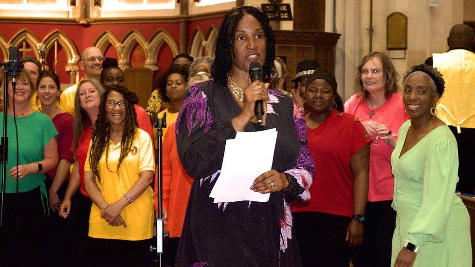 A woman gives a speech holding a microphone while a choir watches on in the background