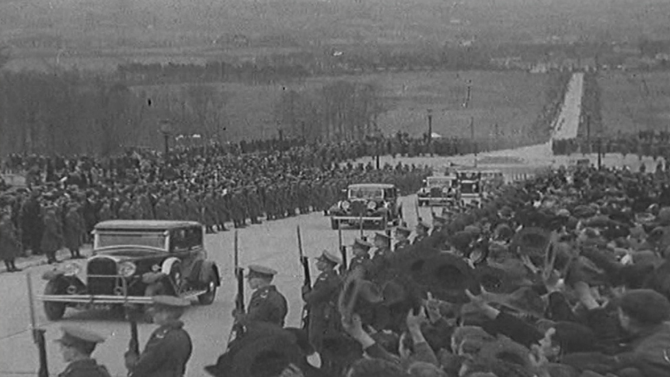 Crowds of people watch as cars pass them on the road to Parliament Buildings at Stormont in the 1930s