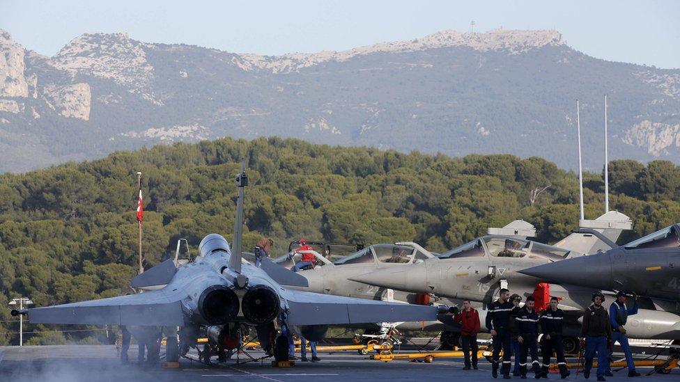 French Navy crew members walk past Rafale fighter jets aboard the French nuclear-powered aircraft carrier Charles de Gaulle before its departure