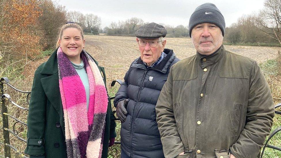 Three people standing in front of the land in winter coats - one wearing a thick colourful scarf. The land is showing it is winter, with leaves off the trees and the field bare.