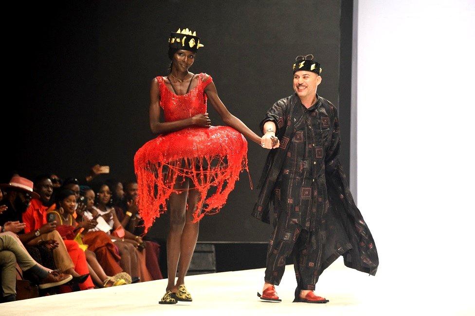 A statuesque model wearing an intricate red dress walks on the runway next to the designer.