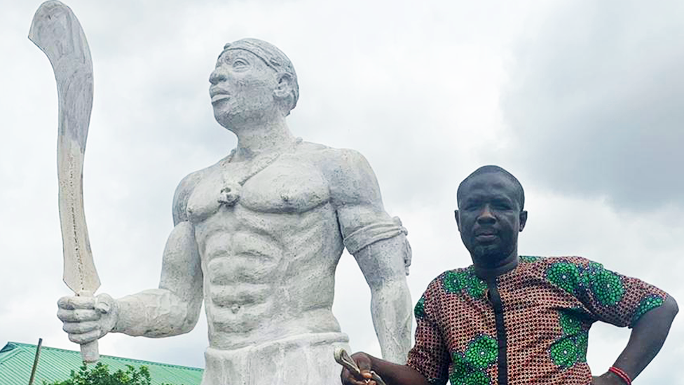 Monday Aigbe standing alongside a statue of his great-grandfather