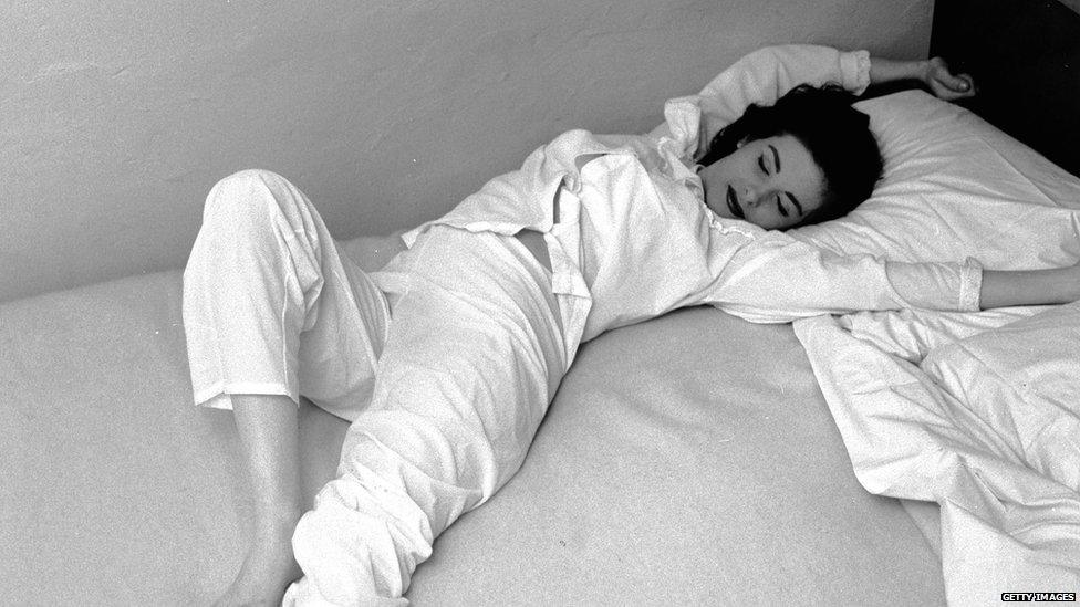 Black and white photo of a woman in pyjamas stretching on a bed