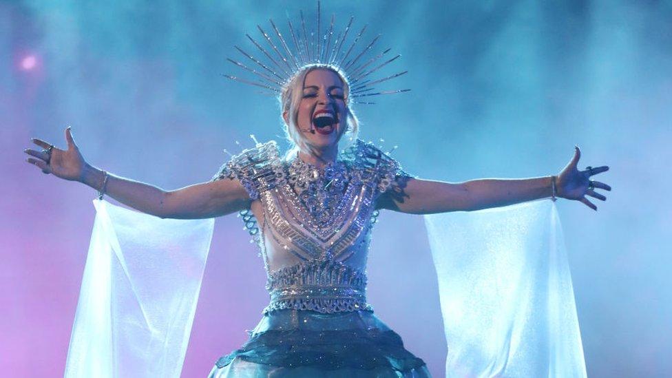 A singer with an elaborate wired headpiece and floaty material coming from her arms sings out with her arms outstretched