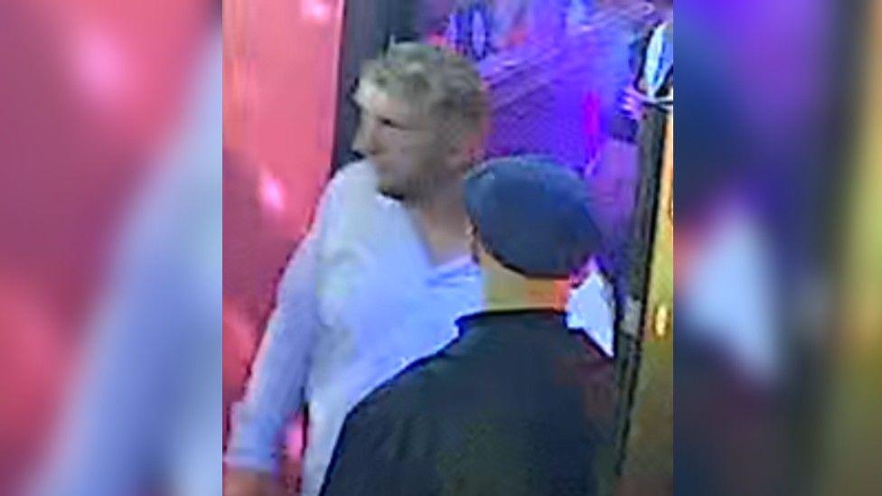 Blurry CCTV image of two middle aged men - one is wearing a black jacket and flat cap with his back to the camera, and the other is facing towards the camera, with shaggy but short greying blond hair and a blue long-sleeved shirt with the top button undone.