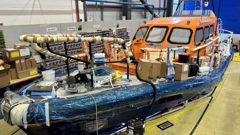 The lifeboat inside a warehouse. It has an orange cabin and a navy hull. It is covered in various tape, sheets and boxes as it is under construction. 