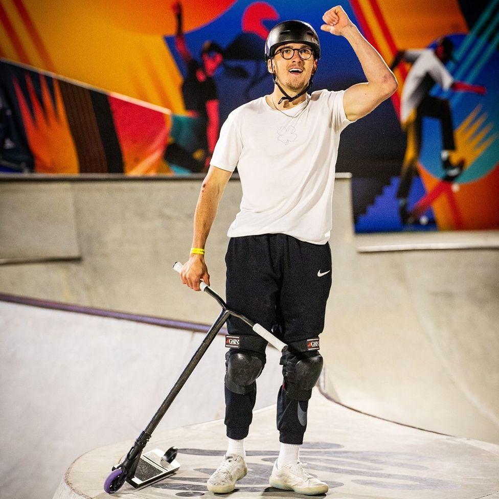 Jayden Sharman holding his left arm in the air at a skatepark in Italy. He is holding his scooter to his right hand side. He is wearing a white T-shirt and black tracksuit bottoms. He has a helmet and knee pads on.