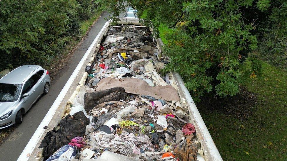 A huge trailer of rubbish. We can see it has been dumped in the middle of a road. It is taking up most of the space, there is a silver car that can't fit on to the remaining sliver of road. We can see a tree that is to the other side of the trailer and hanging over. Inside the trailer is a selection of rubbish including bin bags and other rubbish-like debris. 