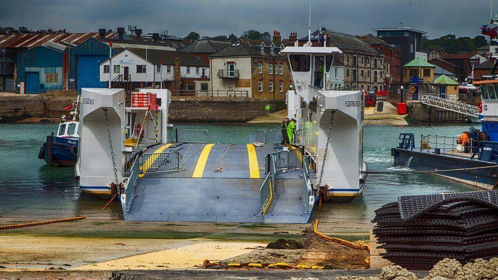 Slipway leading to waiting chain ferry with its ramp down and buildings in the background on the other side of the River Medina