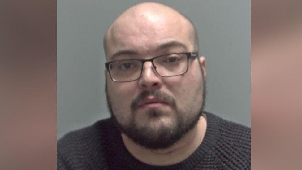 Geddes Abbott a man who is looking directly at the camera and is wearing a dark grey sweater and glasses.