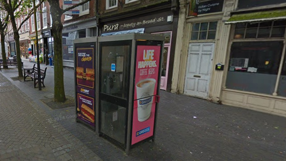 Two grey phone boxes are next to each other on Broad Street in Worcester. They are on a city street with shops in the background as well as two benches and trees to the left