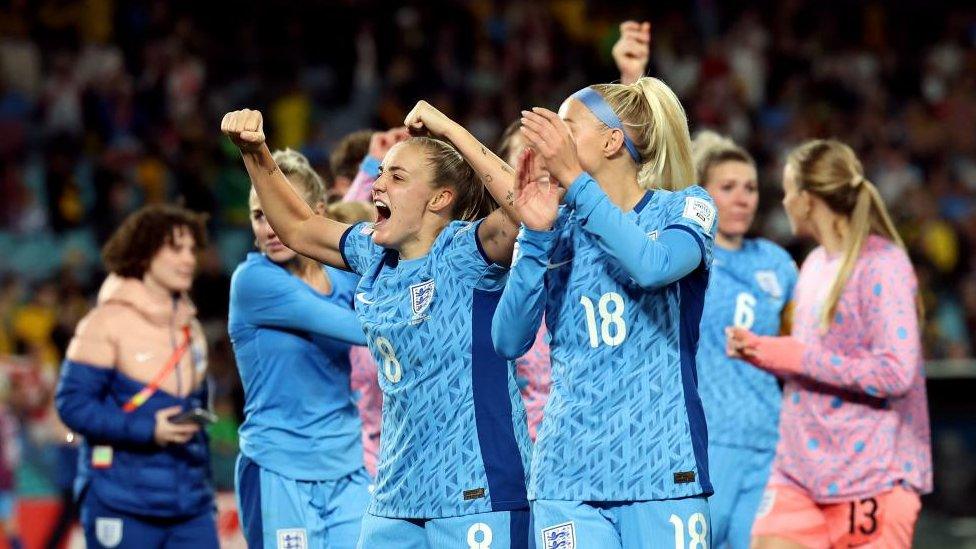 England's Georgia Stanway and other members of the team celebrate beating Australia to progress to the final of the women's World Cup. Stanway is holding both fists in the air. Another player in the foreground claps.