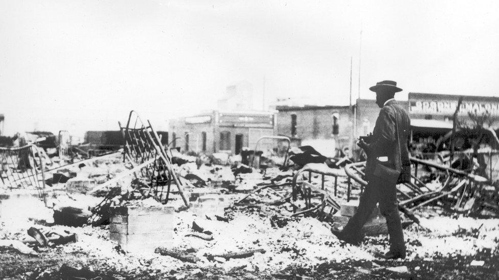 Photograph of an African-American man with a camera looking at the skeletons of iron beds which rise above the ashes of a burned-out block after the Tulsa Race Riot, Tulsa, Oklahoma, 1921