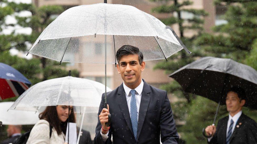 Rishi Sunak walks towards the camera during his visit to the G7 summit in Hiroshima, Japan. He's holding a clear plastic umbrella, dotted with raindrops. There are other people holding umbrellas in the background