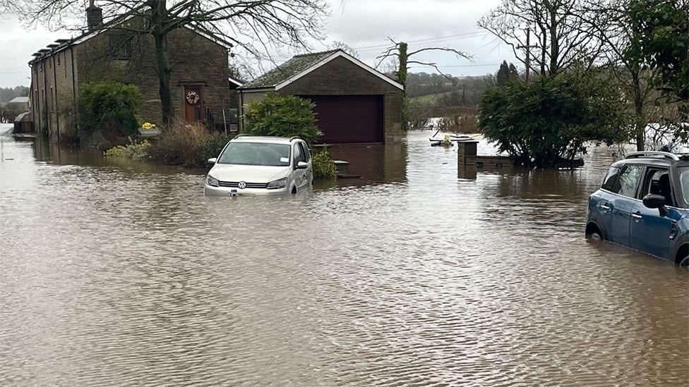 A row of terraced houses are surrounded by brown muddy water, with a grey Vauxhall and blue Mini-Cooper car also partially submerged