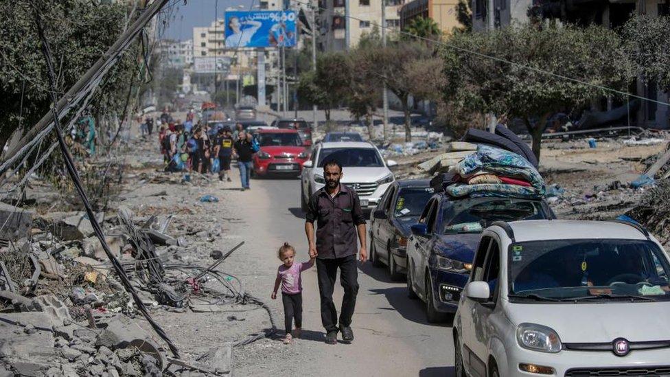 Residents of Gaza City evacuate ahead of an expected ground offensive by the Israeli military. A line of cars is seen on the road. A man in the foreground walks along the side of the road leading a small girl by the hand.