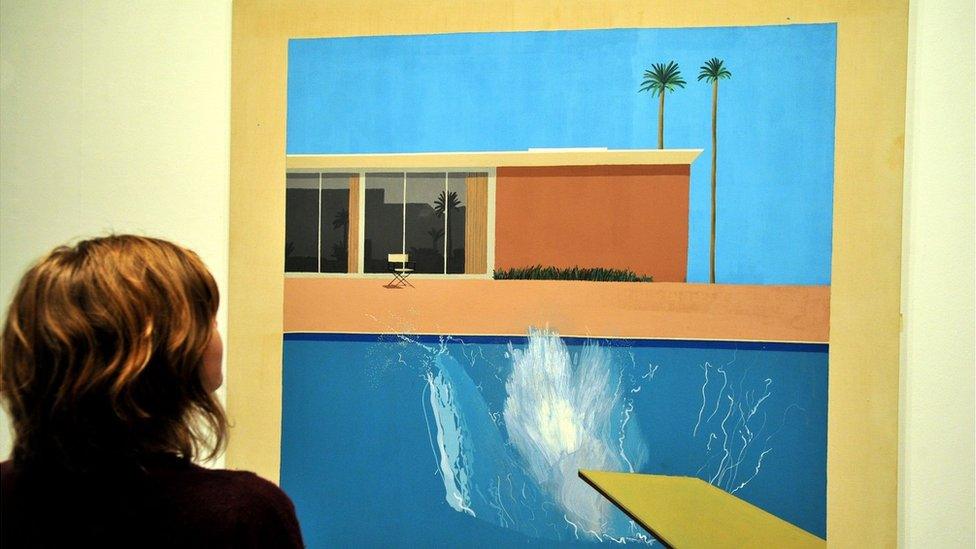 A woman looking at the David Hockney painting A Bigger Splash, It depicts a diving board over a swimming pool, a splash in the water and a building and palm trees against a cloudless blue sky