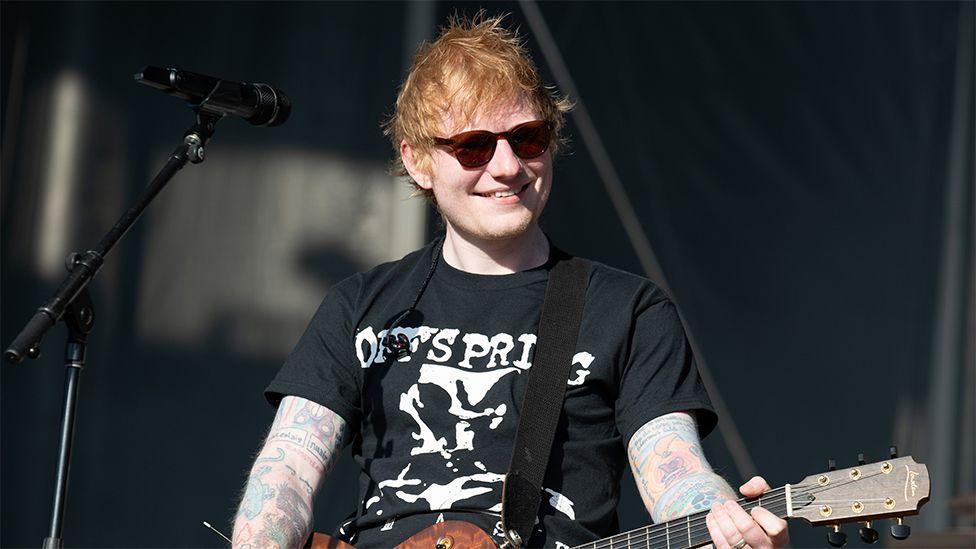 Ed Sheeran, smiling and wearing a black tshirt with white writing, brown sunglasses, performing on stage with a guitar in hand and a microphone on a stand next to him.