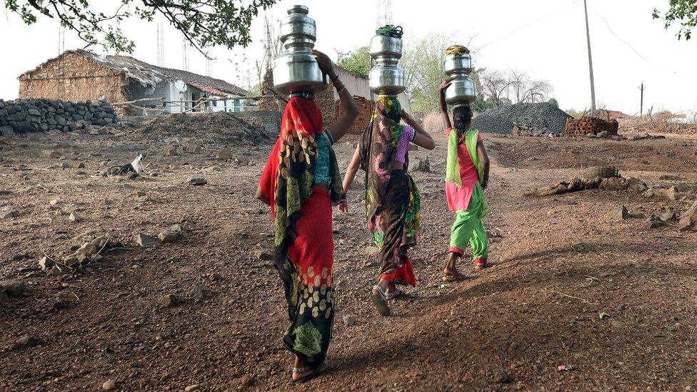 Three women carrying metal containers of water on their heads. They wear brightly coloured clothes.