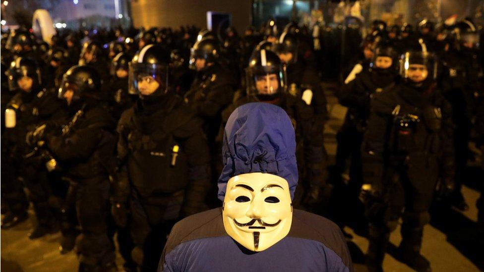 A demonstrator wearing a Guy Fawkes mask on his back stand in front of riot police during a protest outside the headquarters of the Hungarian state television in Budapest (December 17, 2018)