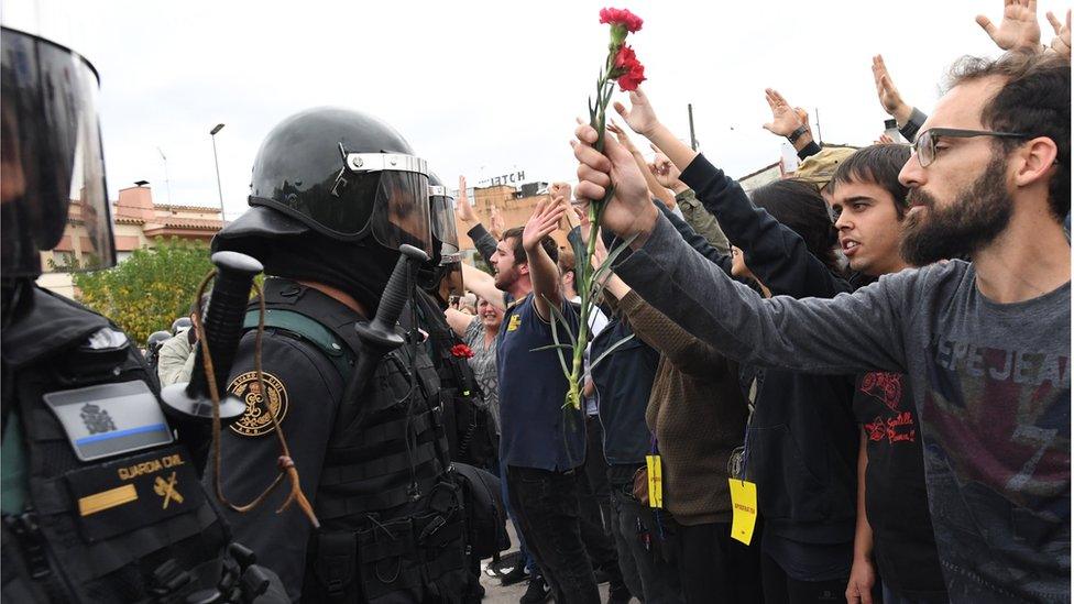 Crowds raise their arms up and a man holds red and pink flowers as police move in on members of the public gathered outside to prevent them from voting in the referendum at a polling station where the Catalonia President Carles Puigdemont will vote later today on October 1, 2017 in Sant Julia de Ramis