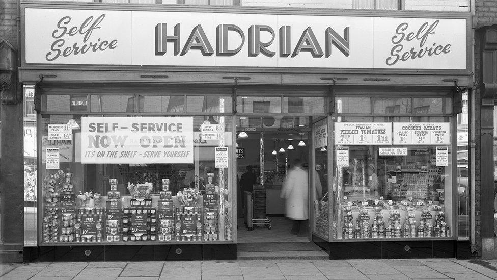 A black and white view of a shop front with lots of signs offering self-service