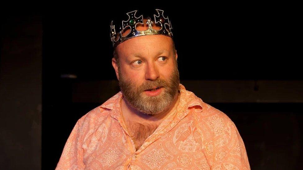 Howard Coggins as Henry VIII wearing a plastic gold crown with a beard and an orange shirt.