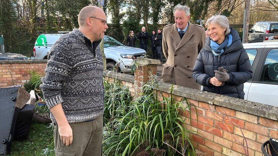 The duchess and a man in a brown coat speaking over a garden wall to Alistair Sinclair