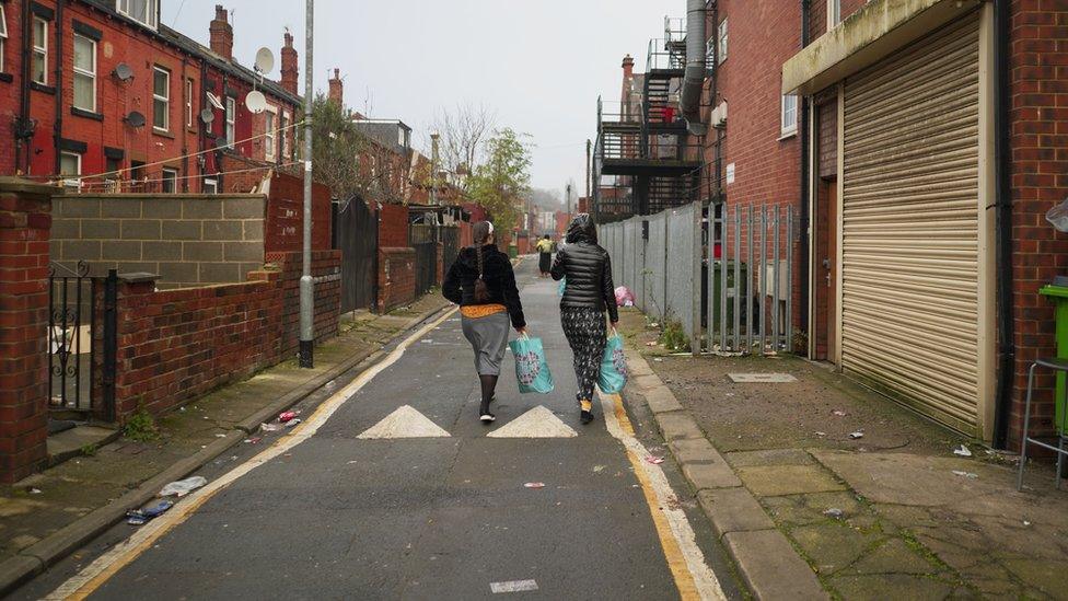 Two women walk down a road holding blue bags full with food