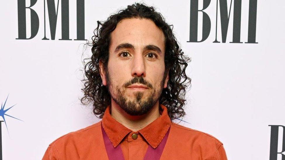 Aaron Horn at the BMI Awards. Aaron has curly dark hair cut just above his shoulders and a short dark beard. He has brown eyes and looks at the camera. He is wearing a burnt orange shirt buttoned to the top with a red silk around his neck which has a medal on it. He is pictured in front of a white back drop with the BMI logo printed on it.