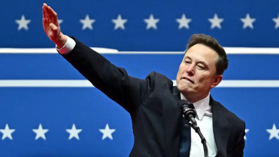 Elon Musk. His arm is extended outwards and up at a 45 degree angle from his body. His hand is mostly flat with the palm down towards the floor.