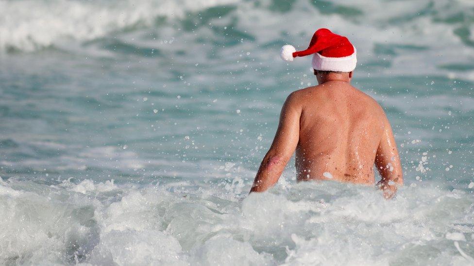 A man swimming in the water ocean. He is wearing a Christmas Santa Claus hat. He is swimming in the waves. The water is blue and white.